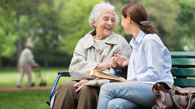 Ältere Frau sitzt mit junger Frau auf einer Bank im Park und beide lächeln sich an | GettyImages / FredFroese