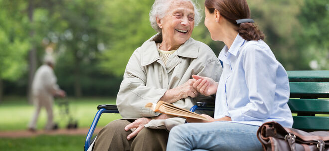 Ältere Frau sitzt mit junger Frau auf einer Bank im Park und beide lächeln sich an | GettyImages / FredFroese