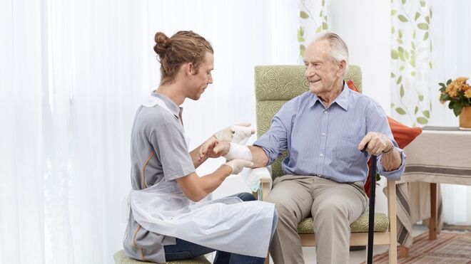 Älterer Mann mit Gehstock in der Hand sitzt lächelnd auf einem Stuhl während ein Pfleger ihm einen Verband umwickelt | FSW